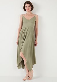 Olive green sleeveless dress with a fitted bodice, A-line silhouette, and asymmetric hem. Made of textured fabric and paired with nude sandals.