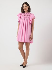 Sieviete stāv rozā ceļgala garuma kleitā ar volāniem un apdari, valkājot melnus šaurus pirmajā debesī aksesuārus ar atvērtu purngalu uz vienkrāsaina fona.