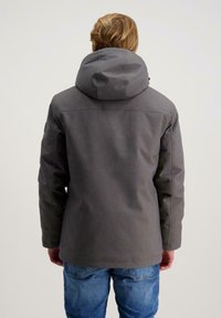 Persoon met lichtbruin haar in een grijze hoodie en blauwe jeans, staande met de rug naar de camera tegen een effen achtergrond.