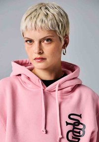 Sudadera rosa con cordones, que presenta un diseño gráfico negro en el lado izquierdo. La tela parece suave y tiene un corte relajado.