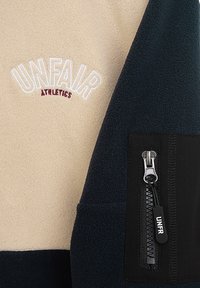 Gyapjúdzseki, amely krémszínű felső résszel és sötétkék alsó résszel rendelkezik. Fekete cipzáras zsebbel és fehér-burgundi "UNFAIR ATHLETICS" hímzéssel készült.