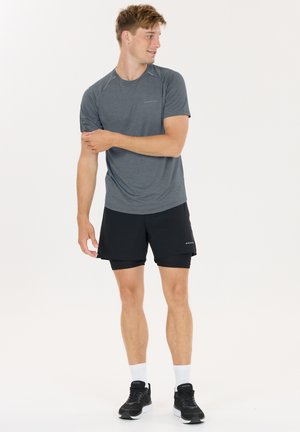 Junger Mann, der den Ärmel eines grauen Sportshirts hochkrempelt, trägt schwarze Lagenshorts, weiße Socken und schwarze Laufschuhe, steht vor weißem Hintergrund.