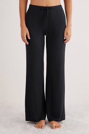 Vrouw staat blootsvoets in zwarte, wijde loungebroek met een elastische tailleband tegen een neutrale achtergrond.
