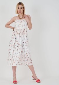 Weißes Blumenkleid mit gestuften Design, versehen mit bunten Blumenmustern. Modell trägt rosa High Heels und eine robuste goldene Halskette.