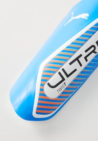 Light blue football shin guard with orange striped pattern, white accents, rounded edges, and "ULTRA TWIST" branding on the front.
