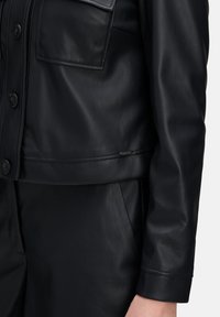 Zwart leren jack met een cropped ontwerp, knoopsluiting aan de voorkant, borstzakken en een gladde textuur met een subtiele glans. Op maat gemaak fit bij de taille.