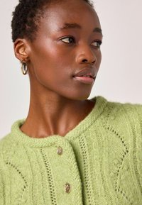 Cardigan verde tricotat, cu un model texturat, guler rotund și două nasturi aurii în față. Material moale, pufos.