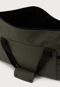 Mørkegrønn stor totebag med svarte håndtak og åpen toppglidelås, som viser svart innvendig fôr.