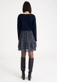 Granatowy dziergany sweter na warstwowej sukience w kwiaty w niebieskie, białe i burgundowe wzory; połączony z czarnymi kozakami do kolan.