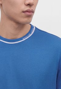 Gros plan d'une personne portant un t-shirt bleu à col rond avec le texte "Michael Kors" sur le col, montrant le bas du visage et le cou.