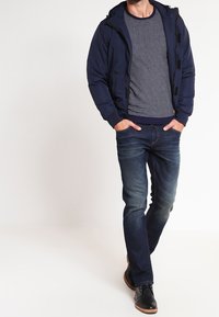 Laivaston sininen hupullinen takki raidallisen neulepuseron päällä, yhdistetty tummansinisiin farkkuihin ja mustiin kenkiin. Takissa on vetoketjullinen suljenta ja sivutaskut.