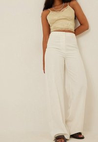 Woman wearing beige lace-trimmed crop top, high-waisted wide-leg white pants, layered gold necklaces, and brown sandals against a plain background.