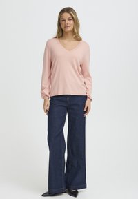 Pull rose clair à col en V avec manches longues, associé à un jean large bleu foncé. Texture lisse et coupe régulière, détails de couture minimaux.