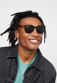 Sorte Ray-Ban solbriller med mørke linser, der har en klassisk rund form og metal accenter, båret med en denimjakke over en teal skjorte.