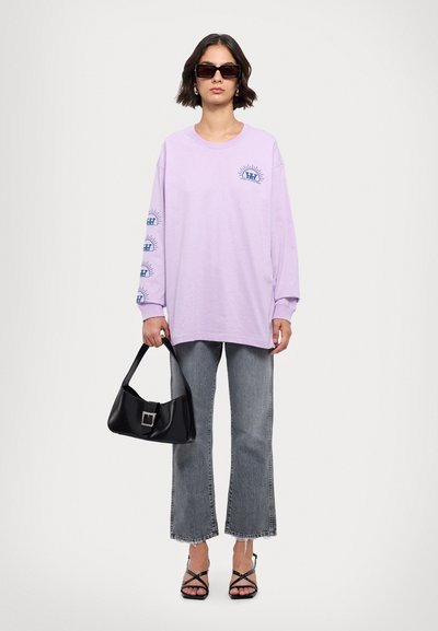 Camiseta de manga larga lila con gráficos en el pecho y las mangas, combinada con jeans de campana grises y un bolso negro. La modelo lleva gafas de sol oscuras.