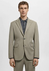 Beige suit jacket with notched lapels, single button closure, two front pockets, and a left chest pocket, worn over a dark polo shirt.