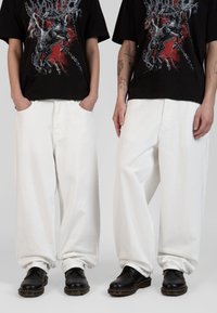 Jeans de mezclilla anchos y blancos con una textura suave, combinados con zapatos de cuero negros. El modelo lleva una camiseta gráfica negra, con las manos en los bolsillos.