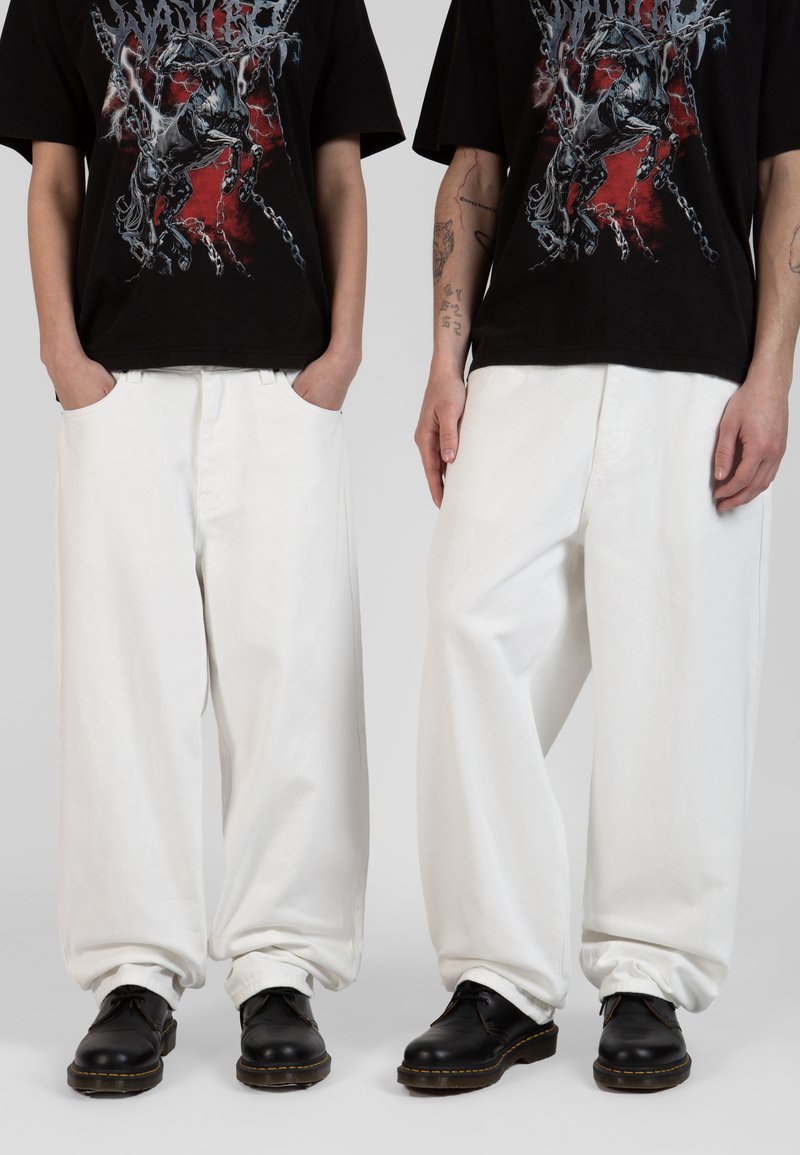 Jeans de mezclilla anchos y blancos con una textura suave, combinados con zapatos de cuero negros. El modelo lleva una camiseta gráfica negra, con las manos en los bolsillos.