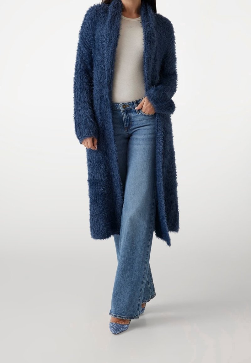 Femme portant un long cardigan bleu duveteux, un haut blanc, un jean bleu à jambes larges et des talons bleus à bout pointu avec des brides à la cheville, debout avec une main dans la poche.
