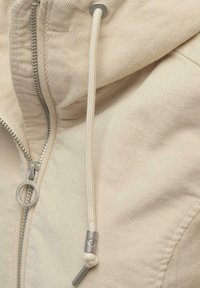 Beige manchesterjacka med en silverfärgad dragkedja med ringdrag, en justerbar huva med vit dragsko och mjukt texturerat tyg.