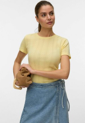 Woman with pulled-back hair wearing a yellow short-sleeve top and denim wrap skirt, holding a brown soft clutch bag with a wooden handle.