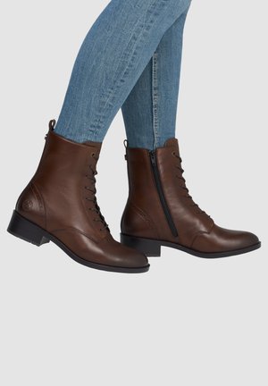 Brown leather lace-up ankle boots with side zipper worn with blue skinny jeans against a plain background.