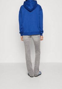 Felpa blu con cappuccio e vestibilità rilassata, abbinata a jeans grigi chiari e sandali a strisce blu, vista da dietro.