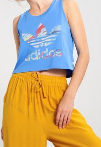 Blå kortärmad tanktopp med en färgglad Adidas-logotyp; har rund hals och är utan ärmar, kombinerad med gula byxor med elastisk midja.