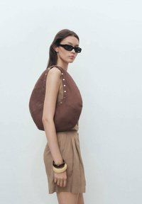 Hnědá látková ramenní taška s odvážným kruhovým tvarem, stříbrné hrotové akcenty, spárována s béžovým outfitem. Modelka nosí tmavé sluneční brýle a náramky.