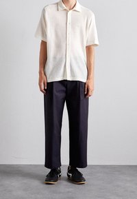 Camisa blanca de manga corta, con un patrón texturizado, combinada con pantalones oscuros de corte holgado y zapatillas deportivas negras.