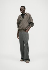 Brun zip-up hoodie, sort t-shirt, grå stribede bredde ben bukser med blomsterbroderi, og lyst fodtøj. Accessories inkluderer solbriller.