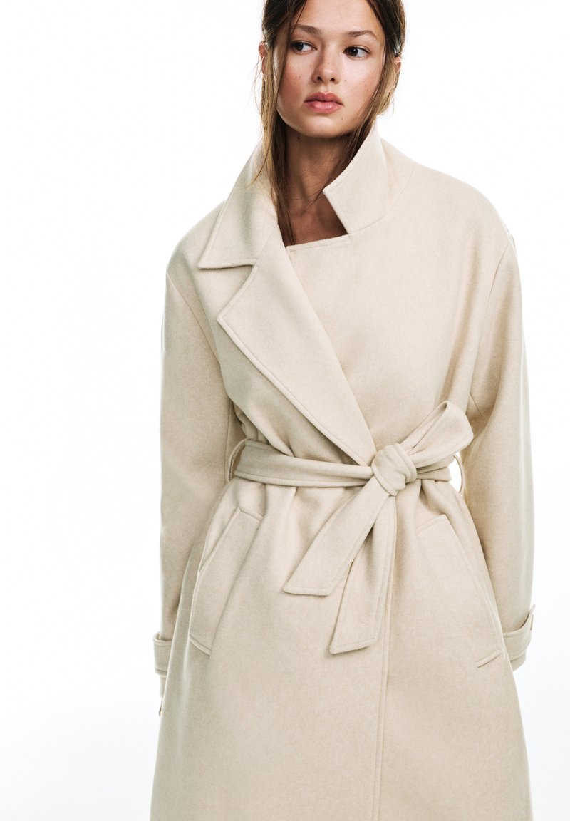 Manteau enveloppant en mélange de laine beige avec un large col, une taille nouée et des poches latérales. Texture lisse et coupe longueur genou.