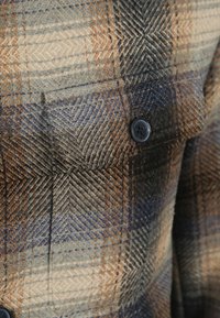 Geruite shirt met tinten bruin, blauw en beige. Zachte textuur met een knoop en een gedetailleerde zak aan de voorkant. Geometrisch patroon.