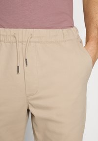 Pantalon beige à cordon de serrage avec un tissu texturé, doté de poches latérales et d'une taille élastique, partiellement visible sous une chemise rose.