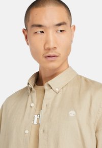Beige linnen overhemd met kraag en knoopsluiting, met een klein geborduurd logo op de borst en daaroverheen een lichtbeige t-shirt.