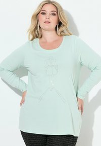 Blondynka z długimi kolczykami nosi jasnymietową, długorękawową koszulę z konturem żakietowej sukienki z kryształków na przodzie.