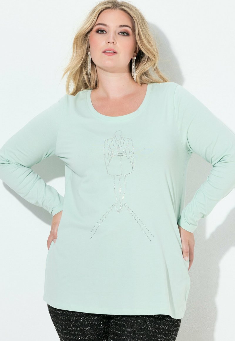 Blondynka z długimi kolczykami nosi jasnymietową, długorękawową koszulę z konturem żakietowej sukienki z kryształków na przodzie.