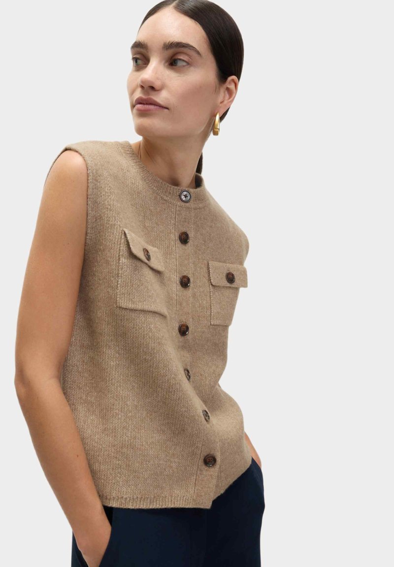 Gebreid mouwloos topje in beige, met een ronde hals, waarbij de voorkant knopen heeft en twee borstzakken met knoopkleppen. Zachte textuur en een getailleerde pasvorm.