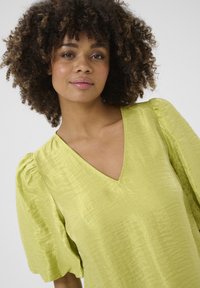 Lime zaļš blūze ar V-veida kakla izgriezumu, papildināta ar pūkainām piedurknēm un teksturētu audumu, kura virsma atstaro gaismu ar maigu spīdumu.