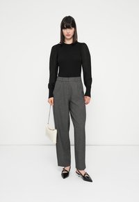 Zwarte gebreide top met getextureerde schouderaccenten, gecombineerd met hoogtaille grijze geruite broek. Model houdt een witte handtas vast; zwarte puntige flats.