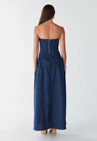 Tmavomodré maxi šaty bez ramienok s priliehavým živôtikom a rozšírenou sukňou, so zlatým zipsom na zadnej strane a jemnou, plynulou textúrou.