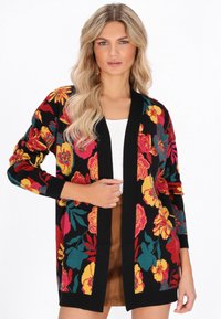 Donna bionda che indossa un cardigan nero con grandi motivi floreali colorati, una maglietta bianca e una gonna marrone, in posa con una mano vicino al bordo del cardigan.