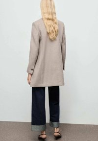 Manteau en laine gris clair avec une coupe droite, un bouton unique et des poignets bas, assorti à un pantalon en denim large de couleur foncée et des sandales marron.