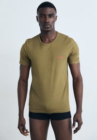 T-shirt vert olive à manches courtes en coton, avec un col rond et un logo "HUGO" rose sur la poitrine. Design simple et ajusté.
