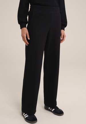 Frau trägt schwarze, strukturierte Marlenehosen und schwarze Sneaker mit weißen Streifen, steht vor einem neutralen Hintergrund.