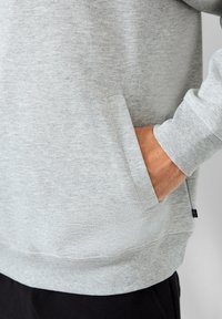 La mano di una persona è appoggiata all'interno della tasca laterale di una felpa grigia chiara con polsini a costine, mentre sono visibili dei pantaloni neri.