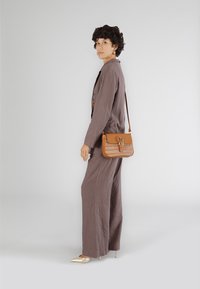 Tutina in lino marrone con gambe ampie, abbinata a una borsa a tracolla beige con un frontale intrecciato e un dettaglio a fibbia. Tacchi a punta dorati.