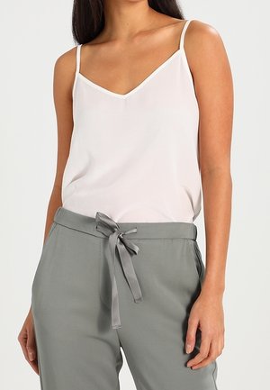 Femme portant un haut blanc sans manches en V et un pantalon gris noué à la taille, debout devant un fond uni.