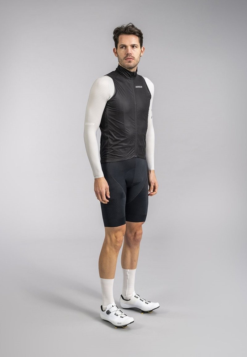 Zwarte mouwloze fietsvest over een witte lange mouw shirt, gecombineerd met zwarte gewatteerde shorts en witte fietsschoenen met zwarte accenten.