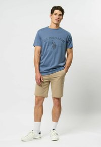 Mėlyna marškinėliai su U.S. Polo Assn. logotipu, derinami su rusvomis šortais. Baltos sportinės bateliai ir trumpi kojinės. Kasdieniška apranga, medvilnės tekstūra.